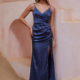 A woman stands on light-colored steps in a sophisticated setting, wearing a deep blue NEPTUNE TO2426 satin, floor-length gown with spaghetti straps, a ruched side, and a thigh-high slit. With one hand on her hip and an elegant, poised expression on her face, she perfectly showcases the style. Angels Formal Wear