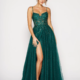 A woman stands confidently in the stunning NIARA PO2488 emerald-green gown, featuring spaghetti straps, intricate beadwork, and a high slit on one side. The dress flows elegantly to the floor, paired with nude heels. Her hair is styled in a chic updo with soft bangs. Angels Formal Wear