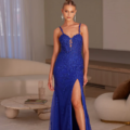 A woman stands in an elegant room, wearing the blue, sleeveless PAVATI PO2487 gown adorned with lace details and a thigh-high slit. She poses with one hand on her hip. Her hair is styled in an updo and she wears long, dangling earrings. The room is decorated with modern, minimalist decor. Angels Formal Wear