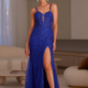 A woman stands confidently in a stylish living room, wearing the elegant PAVATI PO2487 gown, a deep blue floor-length dress adorned with floral embroidery and featuring a thigh-high slit. Her hair is styled up, and she accessorizes with statement earrings and open-toed heels. The sophisticated setting showcases white furniture in the background. Angels Formal Wear