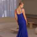 A woman stands with her back to the camera, wearing a floor-length, royal blue, lace dress with thin straps from the PAVATI PO2487 collection. She has her blonde hair styled in an elegant bun. The room features sheer white curtains, a round rug, a circular coffee table, and neutral-toned furniture. Angels Formal Wear