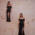 Two women dressed in off-shoulder black RIALTA TO2425 dresses with thigh-high slits are positioned in front of a textured beige wall adorned with symmetrical stepped platforms. The woman on the left stands on an elevated platform, while the woman on the right remains at ground level, exuding elegance and sophistication. Angels Formal Wear