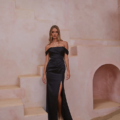 A woman stands on light stone steps against a pale pink wall, wearing an off-the-shoulder, form-fitting black dress with a thigh-high slit. Her long, wavy blonde hair drapes over her shoulders. A small archway and the RIALTA TO2425 product are visible in the background. Angels Formal Wear