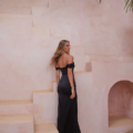 A woman with long hair is depicted from behind, standing on stone stairs in a sandy-colored, minimalistic setting featuring an arched doorway. She is dressed in an off-the-shoulder, floor-length black gown by RIALTA TO2425. Palm leaves are visible in the top corners of the image. Angels Formal Wear