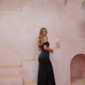 A woman with long, wavy hair wearing an off-the-shoulder, form-fitting black dress stands on a set of light pink, stucco stairs, looking back over her shoulder. The background includes a curved arch and some palm fronds swaying in the breeze exemplifying the elegance of the RIALTA TO2425 setting. Angels Formal Wear