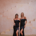 Two women in matching black RIALTA TO2425 off-the-shoulder dresses, each with a thigh-high slit, pose on a sandy floor in front of a geometric, burnished pink wall with step-like structures. They stand next to each other with relaxed and confident expressions. Angels Formal Wear