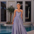 A woman stands on a patio in front of a pool, donning the stunning SEINE PO2490, a strapless lavender evening gown with elegant draping and embellishments. She has her hair styled in an updo and is smiling. A tall plant graces the left side, with modern architecture providing a striking backdrop. Angels Formal Wear