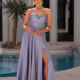 A woman with her hair in a bun is posing outdoors in front of a pool, wearing the SEINE PO2490 lavender dress. The dress features floral embroidery on the bodice and a high slit on one side. She is smiling, with a modern building and a desert plant in the background, enhancing her elegant look. Angels Formal Wear