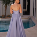 A woman with blonde hair in a bun stands with her back to the camera. She is wearing a strapless, floor-length lavender gown with a fitted bodice, unmistakably reflecting the elegance of SEINE PO2490. She is near a swimming pool, and the ambiance is enriched by a small palm tree in the background, capturing an outdoor idyll. Angels Formal Wear