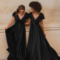 Two women in flowing black AUCKLAND TO872 dresses stand side by side against a textured beige wall in Auckland. They smile and hold hands, with one woman gracefully lifting her dress to display its elegant drape. Both dresses have V-necklines and cap sleeves. Angels Formal Wear