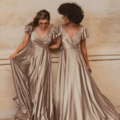 Two women stand side by side in floor-length, metallic gold dresses named AUCKLAND TO872, featuring flutter sleeves and V-necklines. Both smile while gazing downward, gently holding the skirts of their dresses. The backdrop is a simple, lightly textured beige wall that echoes the elegance seen at Auckland TO872 events. Angels Formal Wear