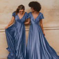 Two women wearing matching AUCKLAND TO872 blue floor-length dresses stand close to each other in a picturesque scene that feels right out of Auckland. Both dresses feature short, flutter sleeves and deep V-necklines. Smiling and looking at each other, they hold their long skirts against a textured, neutral-toned wall. Angels Formal Wear