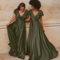 Two women stand side by side in elegant, flowing, dark green AUCKLAND TO872 gowns with V-necklines and flutter sleeves. Both are smiling and looking at each other, each gently holding the skirt of their dress. They are in front of a beige, textured wall. Angels Formal Wear