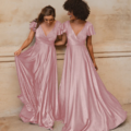 Two women in elegant AUCKLAND TO872 pink dresses stand side by side, smiling and holding the flowing skirts of their dresses. They are in front of a textured beige wall in Auckland, creating a warm and joyful atmosphere. Angels Formal Wear