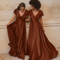 Two women wearing matching floor-length, rust-colored silk dresses with short flutter sleeves and V-necklines stand against a beige textured wall. In Auckland, one lifts the side of her dress while the other touches her hair, both smiling. Their elegant attire radiates AUCKLAND TO872 quality. Angels Formal Wear