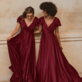 Two women stand side by side in Auckland, each wearing a long, deep red, flowing dress with short ruffled sleeves. They smile at each other against a neutral-toned wall, one holding up the hem of her AUCKLAND TO872 dress. Angels Formal Wear