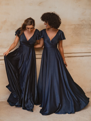 Two women stand together wearing elegant, floor-length AUCKLAND TO872 gowns in navy blue, featuring deep V-necklines and flutter sleeves. One woman holds the side of her dress as they both smile and look at each other against a neutral-colored backdrop reminiscent of an Auckland evening. Angels Formal Wear