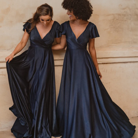 Two women stand together wearing elegant, floor-length AUCKLAND TO872 gowns in navy blue, featuring deep V-necklines and flutter sleeves. One woman holds the side of her dress as they both smile and look at each other against a neutral-colored backdrop reminiscent of an Auckland evening. Angels Formal Wear