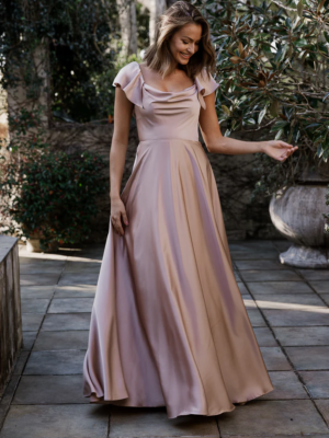 A woman stands outdoors on a stone pathway, adorned in a flowing, light pink, off-the-shoulder LISETTE TO892 gown. She touches the hem of her dress with one hand, smiling and looking down. The background features lush greenery and a large planter. Angels Formal Wear