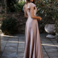 A woman stands on a stone-paved area enveloped by lush greenery and large planters. She is dressed in a flowing, floor-length light mauve gown featuring ruffled sleeves and a V-back. As she gazes over her shoulder towards the camera, the serene outdoor setting captures the elegance of LISETTE's TO892 collection beautifully. Angels Formal Wear