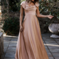 A woman wearing the LISETTE TO892, a flowing peach-colored gown with short, ruffled sleeves, stands outdoors on a stone pathway. She smiles softly, looking down while holding the edge of her dress. Lush greenery and a large planter are visible in the background. Angels Formal Wear