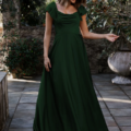 A woman stands outdoors on a tiled patio, wearing a floor-length, dark green LISETTE TO892 dress with short flutter sleeves. She is smiling, holding the side of her dress in one hand, with greenery and trees in the background. Angels Formal Wear