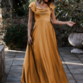 A woman stands outdoors on a stone pathway wearing the LISETTE TO892 gown, a flowing, mustard-yellow dress with cap sleeves and a draped neckline. She looks down and to the side, gently holding a part of her attire. Surrounded by trees and greenery, it's as if she’s stepped straight out of an LISETTE dream. Angels Formal Wear