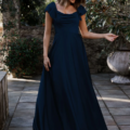 A woman in a navy blue, floor-length LISETTE TO892 dress with short, cap sleeves is standing outdoors. She is holding the hem of her dress with one hand, looking down and smiling. The background features greenery and a stone patio that complements the LISETTE TO892 beautifully. Angels Formal Wear