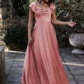 A woman stands outdoors on a stone pathway, wearing the LISETTE TO892, a flowing pink gown with short ruffled sleeves and a draped neckline. She has light hair and smiles while slightly looking downward. The background features greenery and a large stone planter, evoking the elegance of Lisette in the serene setting. Angels Formal Wear