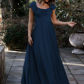 A woman in a LISETTE TO892, a dark blue, floor-length dress with cap sleeves, stands outdoors on a stone patio. She is smiling and lightly holding one hand up. Lisette is surrounded by greenery and stone structures in a sunlit setting. Angels Formal Wear