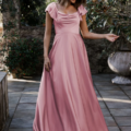 A woman in a flowing pink dress with flutter sleeves stands on a stone patio. She smiles while looking down, holding the edge of her LISETTE TO892 dress with her left hand. The background features greenery and a decorative stone planter. Angels Formal Wear