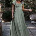 A woman stands outdoors on a stone patio surrounded by greenery. Wearing a LISETTE TO892 light green, floor-length dress with flutter sleeves and a draped neckline, she smiles and looks down to her right, holding part of her dress in her hand. Angels Formal Wear