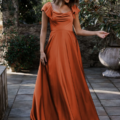 A woman stands outdoors in a shaded patio area, wearing the elegant LISETTE TO892 dress—a flowing burnt orange piece with short sleeves and a fitted bodice. She smiles while looking down and gently holding a part of her dress. Lush greenery is visible in the background. Angels Formal Wear