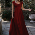 A woman stands in a stone-paved garden, adorned in a flowing, floor-length LISETTE TO892 gown in rich burgundy, featuring elegant short ruffled sleeves. Smiling softly as she looks down, her hand gently caresses the fabric of her dress. The backdrop showcases lush greenery and large decorative pots. Angels Formal Wear