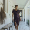 A woman in a shimmering, dark-colored, asymmetrical dress walks down a covered wooden porch adorned with rustic decorations and benches. The porch is part of a light-colored building labeled YOLANDA MO2351, and the scene is well-lit with natural light. Angels Formal Wear