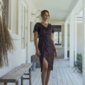 A woman is standing on a wooden porch wearing a dark, shimmering dress with short sleeves and a high slit. She is looking ahead with one hand resting by her side. The porch, part of the YOLANDA MO2351 cabin, has white siding, a bench, and various decorative items hanging on the wall. Angels Formal Wear