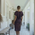 A woman with light brown hair is standing in a brightly-lit hallway with wooden floors, gazing away from the camera. She dons the knee-length, sparkly YOLANDA MO2351 dress with short sleeves and pairs it with light-colored high-heeled shoes. The minimally decorated hallway, bathed in natural light, enhances her serene elegance. Angels Formal Wear