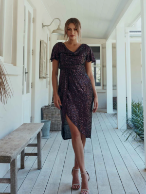 Yolanda stands confidently in a hallway, dressed in the YOLANDA MO2351—a dark, shimmering short-sleeve dress featuring an asymmetrical hemline. Her light brown hair is styled in a loose updo, and she completes her look with strappy high-heeled sandals. The setting appears to be a bright, airy porch. Angels Formal Wear