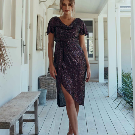 Yolanda stands confidently in a hallway, dressed in the YOLANDA MO2351—a dark, shimmering short-sleeve dress featuring an asymmetrical hemline. Her light brown hair is styled in a loose updo, and she completes her look with strappy high-heeled sandals. The setting appears to be a bright, airy porch. Angels Formal Wear