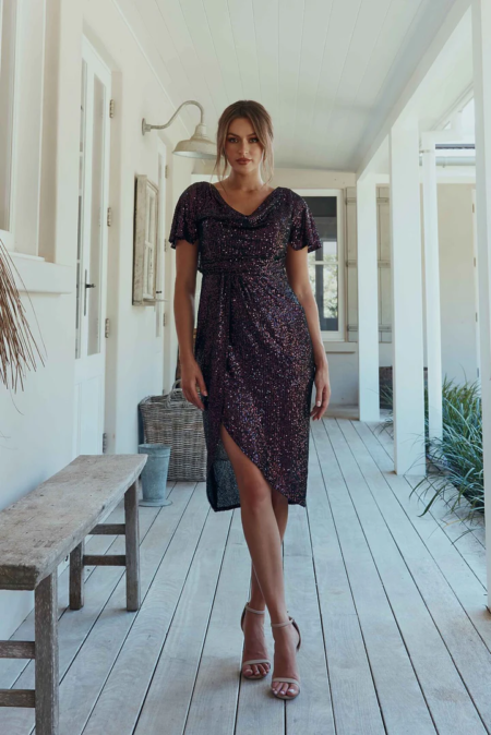 Yolanda stands confidently in a hallway, dressed in the YOLANDA MO2351—a dark, shimmering short-sleeve dress featuring an asymmetrical hemline. Her light brown hair is styled in a loose updo, and she completes her look with strappy high-heeled sandals. The setting appears to be a bright, airy porch. Angels Formal Wear