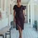 Yolanda stands confidently in a hallway, dressed in the YOLANDA MO2351—a dark, shimmering short-sleeve dress featuring an asymmetrical hemline. Her light brown hair is styled in a loose updo, and she completes her look with strappy high-heeled sandals. The setting appears to be a bright, airy porch. Angels Formal Wear