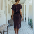 Woman in a YOLANDA MO2351 sequined dress stands on a wooden deck, facing away from the camera. Her hair is tied back, and she is wearing high heels. The deck is decorated with a bench, a basket, and potted plants, leading to a white-walled interior with large windows and doors. Angels Formal Wear