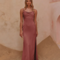 A woman stands on a sandy textured floor, wearing a long, sleeveless, mauve satin dress with a high slit on one side. She has long, blonde wavy hair and is gazing directly at the camera. The background features a light, textured wall with a minimalist archway adorned subtly by the CARY TO2432. Angels Formal Wear