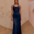 A woman stands on a sand-covered floor against a textured, plaster-like wall. She is elegantly dressed in CARY TO2432, a long navy satin dress with thin straps and a slight cowl neckline. Her long, wavy hair frames her neutral expression as she gazes directly at the camera. Angels Formal Wear