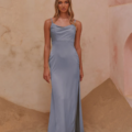 A woman stands in a softly lit setting with sandy and pink-hued textured walls. She is wearing a shimmery, light blue, floor-length gown with thin straps and a draped neckline. Her blonde hair, styled in loose waves, frames her calm expression as she elegantly conceals the CARY TO2432 accessory. Angels Formal Wear