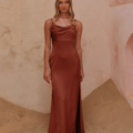 A woman stands on a sandy surface against a textured wall, wearing the sophisticated CARY TO2432 gown. The rust-colored, satin dress features a sleeveless design and a subtle cowl neckline. Her long, wavy hair cascades down her shoulders as she gazes directly at the camera in this elegant shot. Angels Formal Wear