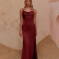 In a room with sandy floors, a woman wearing the satin burgundy CARY TO2432 dress, complete with thin straps, a slight sheen, and a high slit on one side, stands gracefully. Her blonde hair cascades in loose waves as she gazes at the camera, effortlessly embodying the elegance of CARY TO2432. Angels Formal Wear