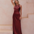 A woman in a sleeveless, burgundy CORDELIA TO2433 satin gown with a fitted bodice and long skirt stands against a light-colored, textured wall. She has one arm raised, resting her hand on her head, while her other hand gently touches the side of her waist. This timeless look evokes the elegance of CORDELIA. Angels Formal Wear