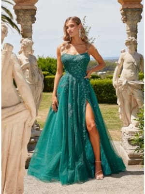 A woman in a teal, floor-length ARALIA PO24155 gown with a high slit stands in an outdoor setting, surrounded by classical statues. The gown features intricate embellishments and she is wearing long, dangling earrings and high-heeled sandals. Angels Formal Wear