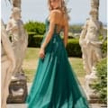 A blonde woman stands outdoors in an elegant setting, wearing the teal, floor-length ARALIA PO24155 gown. The dress features a lace-up back and delicate beadwork. She is surrounded by intricate stone statues and greenery, with a scenic view in the background. Angels Formal Wear